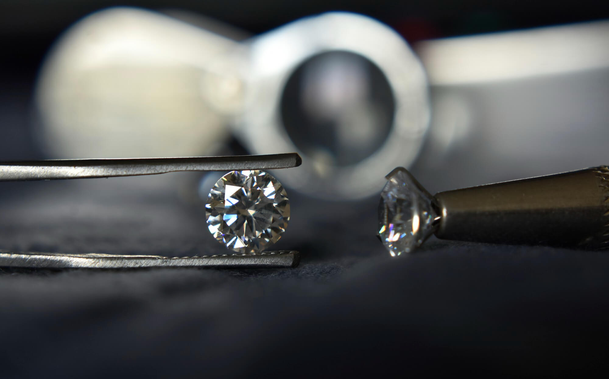 Brilliant cut diamond held in professional tweezers with a jeweler's loupe in the background.
